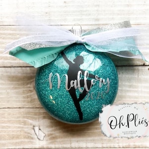 May include: Teal glitter Christmas ornament with a silver glitter silhouette of a ballerina and the name Mallory 2020. The ornament is tied with a white and silver ribbon. The ornament is from Oh.Pliés Dancewear Boutique in Branson, MO.