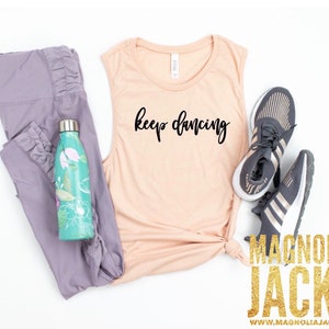 May include: A peach-colored tank top with the text "keep dancing" in black script. The tank top is laid out on a white surface with a pair of gray leggings, a teal water bottle with floral designs, and a pair of gray and gold sneakers.