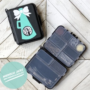 May include: A black plastic storage box with a clear lid, divided into compartments. The box is open and filled with hair accessories, including bobby pins, hair ties, and a hair brush. The box has a graphic of a megaphone with a bow on top and the letters "NFB" inside the megaphone.