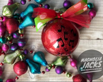May include: Red glitter Christmas ornament with black musical notes and a red and green ribbon. The ornament is surrounded by a garland of colorful ornaments.