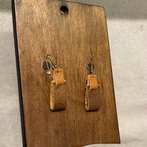 May include: A pair of handmade leather earrings displayed on a wooden stand. The earrings feature a rectangular design with a light brown top and a darker brown bottom. The stand is a dark wood color with a hole at the top.