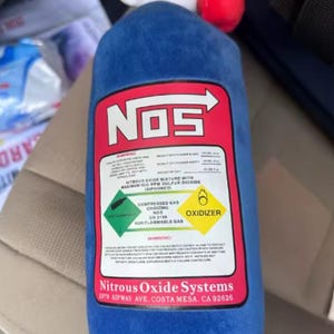 May include: A blue plush toy shaped like a nitrous oxide tank, featuring a red and white label with the "NOS" logo and warning labels. The toy has a white top and a red ball on top. The text on the label includes "Nitrous Oxide Systems".