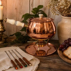May include: A polished copper fondue pot with a white handle and matching lid, resting on a copper stand and base. Four metal fondue forks with wooden handles in orange, red, green, and white are arranged on a white cloth next to the pot.