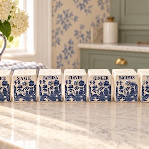 May include: A set of seven square ceramic spice jars featuring a blue and white willow pattern. Each jar is labeled with black text: ALLSPICE, SAGE, PAPRIKA, CLOVES, GINGER, BAYLEAVES, and THYME. The jars have perforated lids for dispensing spices.