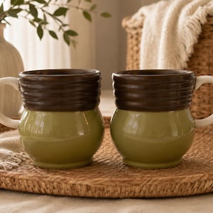 May include: Two ceramic mugs featuring a rounded base in a muted sage green glaze and a dark brown ribbed upper section. Each mug has a smooth, cream-colored handle attached to the side, providing a contrast to the earthy tones of the body.