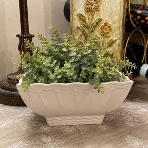 May include: A white ceramic planter with a scalloped rim and a wavy textured base, filled with a lush arrangement of small, round green eucalyptus leaves. The planter has a rectangular shape with vertical ridges along the sides.