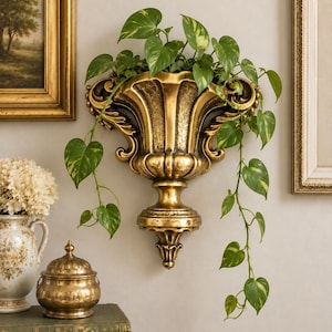 May include: A gold-colored wall-mounted planter with an ornate, fluted design and decorative scrollwork. The planter holds a trailing green pothos plant with variegated heart-shaped leaves that drape down from the top of the vessel.