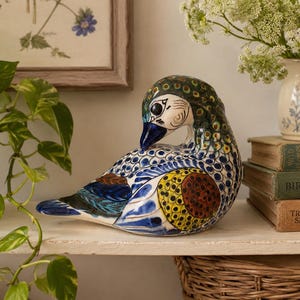 May include: A ceramic duck figurine featuring intricate hand-painted patterns in shades of blue, yellow, green, and brown. The duck is depicted in a preening pose with its head turned toward its back, showcasing detailed plumage designs.