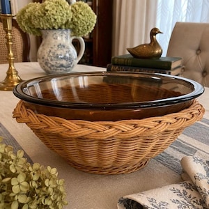 May include: A round, transparent amber-colored glass bowl set inside a natural light brown woven wicker basket holder. The bowl features a smooth, wide rim, while the basket base has a thick, braided edge detail around the top perimeter.