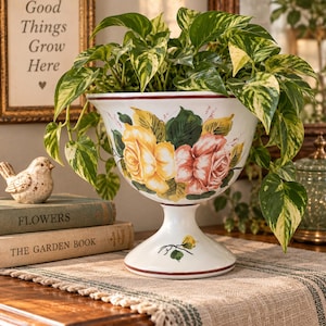 May include: A white ceramic pedestal planter featuring a hand-painted floral design with yellow and pink roses. The bowl-shaped vessel has a flared rim with a thin dark red border and a small painted green leaf motif on the base, holding a pothos plant.