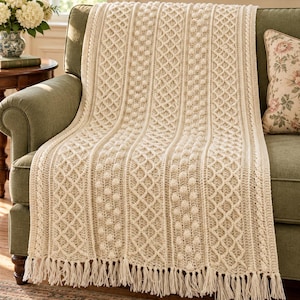 May include: A cream-colored knit throw blanket featuring vertical panels of textured bobbles and diamond-shaped cable patterns. The blanket is finished with long, straight fringe along the bottom edge, creating a detailed and uniform appearance.