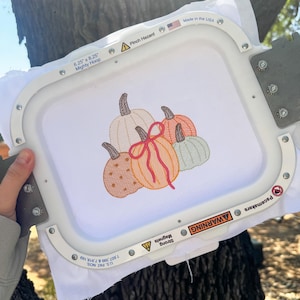 May include: A white fabric square secured in a 15.88 cm by 20.96 cm Mighty Hoop embroidery frame. The fabric features an embroidered design of five pumpkins in shades of cream, orange, brown, and light green, with a bright pink bow tied around the centre pumpkin.