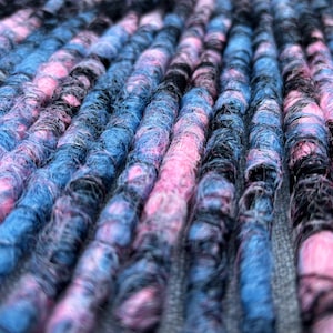 Puede incluir: Primer plano de rastas coloridas en tonos rosa, azul y negro. El cabello sintético está enrollado con fuerza, creando un efecto texturizado y en capas. Las rastas están dispuestas en filas paralelas, mostrando la vibrante mezcla de colores.