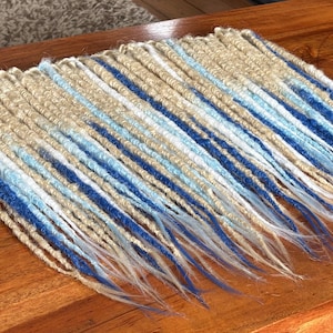 May include: A set of synthetic dreadlocks featuring a color gradient that transitions from a natural beige at the roots to shades of light blue, medium blue, and dark blue at the tips, with thin, textured strands arranged in a parallel formation.