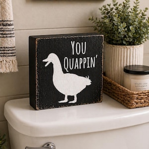 May include: A black wooden block sign with the words "YOU QUAPPIN'" and a white duck silhouette. The sign is placed on a white toilet tank, with a decorative plant and candle in the background.