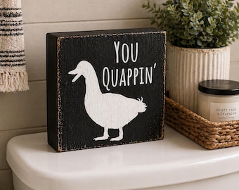 May include: A black wooden block sign with the words "YOU QUAPPIN'" and a white duck silhouette. The sign is placed on a white toilet tank, with a decorative plant and candle in the background.