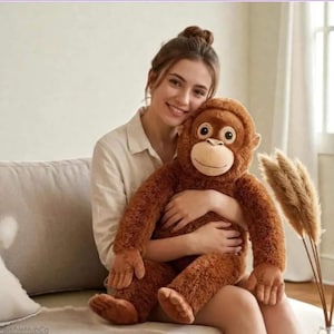 May include: A large, plush brown orangutan stuffed animal with a smiling face and big eyes. The soft toy has long arms and legs, and is being held. The plushie is a cuddly toy.