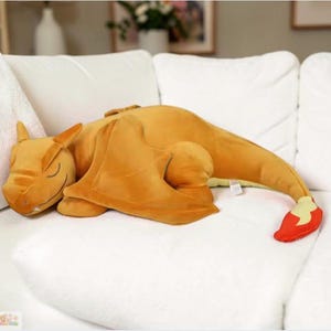 May include: A plush, orange Charizard pillow, lying on a white couch. The pillow features a detailed design with wings, a tail with a red flame, and closed eyes, suggesting a sleeping pose. The plush toy is soft and cuddly.