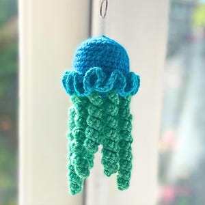May include: A crocheted jellyfish ornament featuring a bright blue rounded head with a ruffled edge and multiple dangling green spiral tentacles. The jellyfish is suspended from a small silver metal ring at the top, showcasing detailed yarn work.