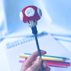 May include: A pen topper featuring a crocheted mushroom design with a red cap and white circular spots, attached to a dark blue pen. The mushroom has a beige stem and is crafted with visible yarn texture, sitting atop a standard writing instrument.