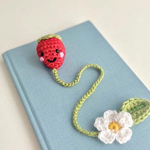 Op de afbeelding: Een handgemaakte gehaakte bladwijzer met een rode aardbei met een lachend gezicht, een groene stengel en een witte bloem met een geel hart. De bladwijzer ligt op een lichtblauw boek.