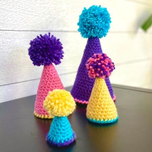 May include: A collection of handmade, colorful crochet party hats with pom-poms. The hats are in various sizes and colors, including pink, purple, yellow, and teal. The pom-poms are in matching or contrasting colors.