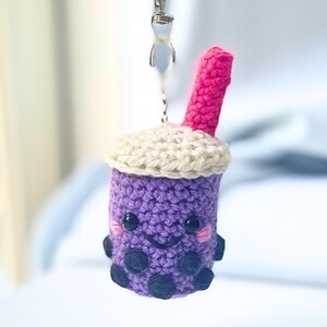 May include: A purple crocheted bubble tea keychain featuring a cream-colored top, a bright pink straw, and black embroidered facial features with pink cheeks. The base is decorated with several small, dark, circular crocheted boba pearls.