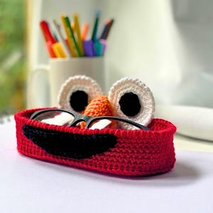 May include: A red crocheted eyeglass holder featuring a black smile, an orange nose, and two white eyes with black pupils. The holder is oval-shaped and contains a pair of black-framed eyeglasses resting inside the structure.