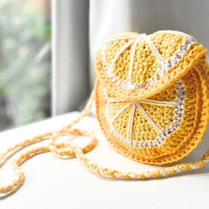 May include: A handmade, crocheted crossbody bag shaped like a lemon slice. The bag is mainly yellow with white detailing, including the strap and the segments of the lemon. The bag is on a white surface.