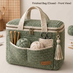 Puede incluir: Un bolso de almacenamiento tejido a ganchillo en color verde salvia con asas color crema, cierre de cremallera y un bolsillo frontal con dos ovillos de lana, tres agujas de madera y tijeras. Una borla crema cuelga de la cremallera y una etiqueta dice "harthuck".