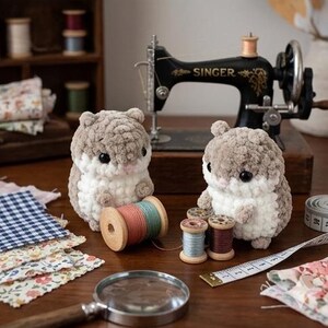 May include: Two plush, crocheted hamster figures with soft, light brown and white yarn textures, black bead eyes, and pink noses. They are positioned near wooden spools of colored thread, a magnifying glass, and a vintage Singer sewing machine.