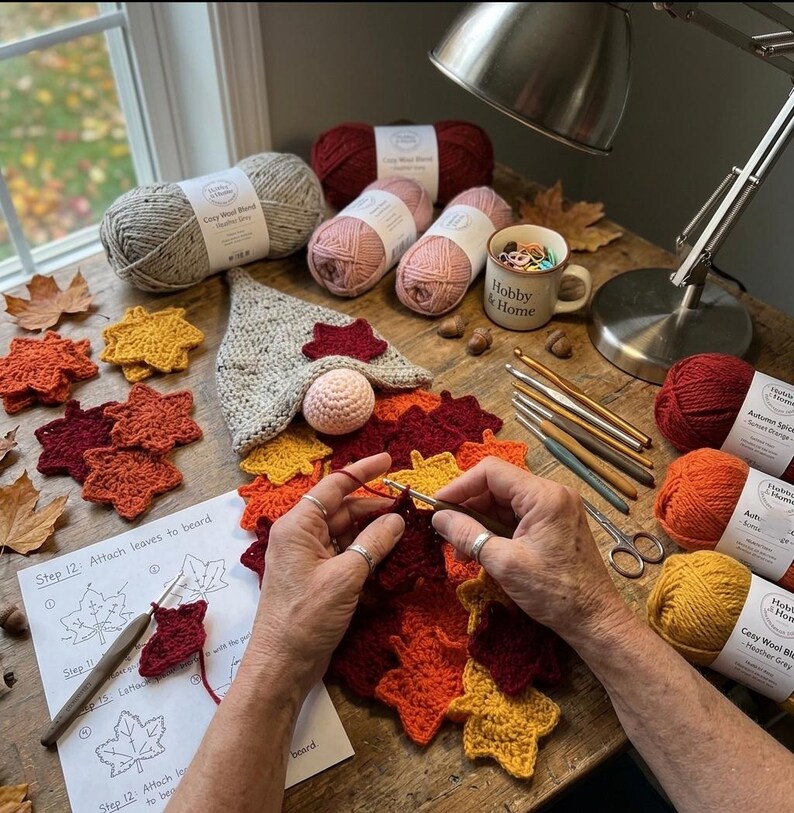 Op de afbeelding: Een haakproject met een kabouter met een gespikkelde grijze muts en een roze neus, omringd door gehaakte herfstbladeren in rood, oranje en geel. Garenbollen, een mok, haaknaalden en een instructieblad liggen op de tafel.