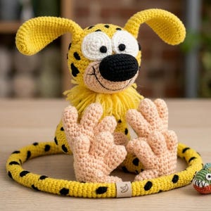 Peut inclure: Un jouet animal en crochet jaune à taches noires avec de grands yeux blancs, un nez noir et un col à franges jaunes. La créature a des mains et des pieds couleur pêche clair, une longue queue tachetée formant un cercle et un petit poisson vert.