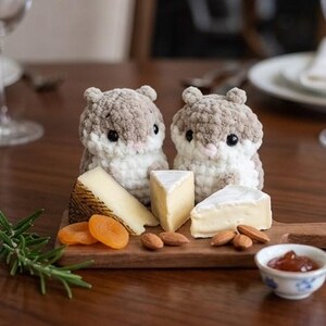 May include: Two crocheted hamster figurines with beige and white yarn, black bead eyes, and pink noses. They are positioned on a wooden serving board alongside wedges of cheese, dried apricots, almonds, a sprig of rosemary, and a small bowl of jam.