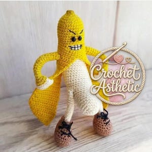 Op de afbeelding: Een handgemaakte gehaakte bananenfiguur met een boos gezicht, gele schil en wit vruchtvlees. De banaan heeft armen, benen en bruine schoenen met zwarte veters. De figuur staat met een cape. De tekst "Crochet Asthetic" staat in een cirkel.