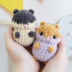 May include: Two small, round, crocheted plush animals resembling hamsters. One is dark gray and cream-colored, while the other is tan and light purple. Both feature black plastic eyes, small pink stitched noses, and textured yarn.