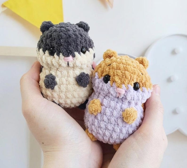 May include: Two small, round, crocheted plush animals resembling hamsters. One is dark gray and cream-colored, while the other is tan and light purple. Both feature black plastic eyes, small pink stitched noses, and textured yarn.