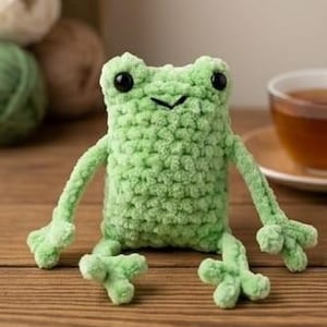 May include: A light green crocheted frog with black eyes and a small, stitched mouth. The frog has outstretched arms and legs, and is sitting on a wooden surface. The frog is a handmade item.