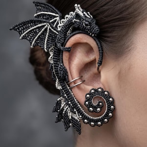 Peut inclure: Un ear cuff dragon noir et argenté avec des détails en cordon tressé, une tête de dragon sculptée sur le dessus, des ailes déployées le long du cartilage de l'oreille et une base en queue en spirale ornée de petites perles argentées.