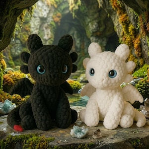 May include: Two plush dragon toys, one black and one white, featuring large blue eyes, textured crochet-style surfaces, small wings, and rounded ears. The black dragon has a small red accent on its tail, and both sit in a seated position.