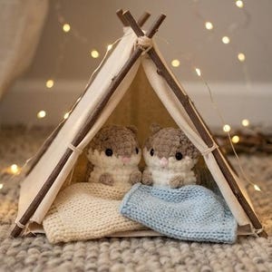 May include: A miniature teepee constructed from wooden sticks and off-white fabric, housing two small, crocheted hamster figures. The hamsters are positioned side-by-side, each tucked under a small knitted blanket, one in cream and one in light blue.