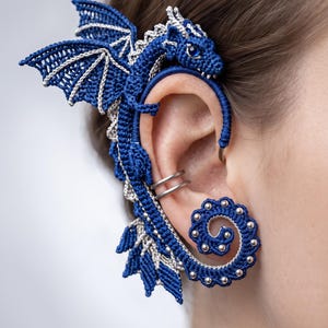 Può includere: Un ear cuff a forma di drago realizzato all'uncinetto in blu e argento, con una testa di drago dettagliata in alto, ali spiegate e una coda a spirale in basso decorata con piccole perline argentate. Il design include due anelli argentati.