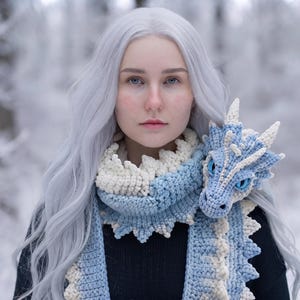 May include: A crochet scarf featuring a light blue and cream dragon design with a detailed dragon head resting on the shoulder. The scarf has a textured, scale-like pattern along the edges and is worn over a plain black long-sleeved garment.