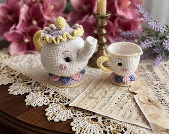 Op de afbeelding: Een gehaakte theepot en bijpassende theekopset in wit met pastelgele, lavendelkleurige en roze accenten. De theepot heeft een deksel met een gele knop, een tuit en een voet versierd met kleine bloemblaadjes. De kop heeft een geel oortje.