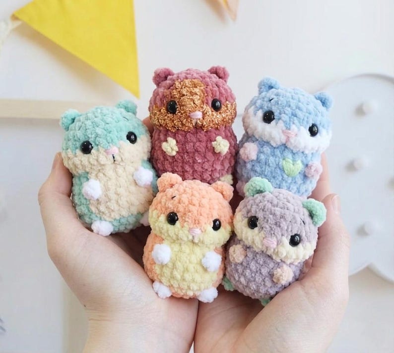 May include: Five small, plush, crocheted hamster figures in pastel and vibrant colors including mint green, dusty rose, light blue, peach, and lavender. Each soft toy features black plastic eyes, a tiny pink nose, and white paws.