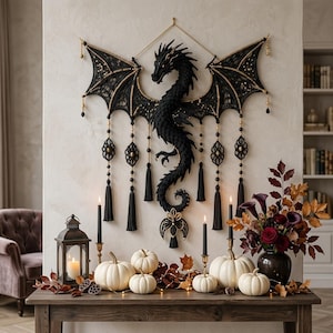 May include: A black macrame dragon wall hanging featuring intricate knotted patterns, gold bead accents, and long black tassels. The dragon has expansive wings and a curved tail, suspended by a gold cord against a light-colored wall surface.