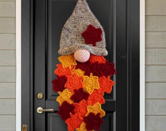 May include: A crochet gnome wall hanging featuring a speckled gray pointed hat adorned with a single dark red leaf. The gnome has a round light pink nose and a long beard constructed from numerous layered crochet leaves in shades of red, orange, and yellow.