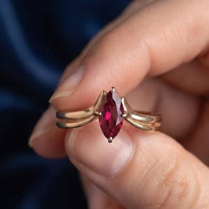 May include: A gold ring featuring a marquise-cut deep red gemstone set in a prong setting. The band has a split design that tapers toward the center stone, creating a sleek and polished look with a vibrant crimson center piece.