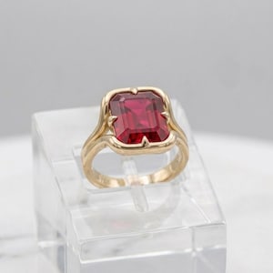 Può includere: Un anello in oro con una pietra preziosa rosso intenso dal taglio rettangolare a smeraldo, montata su quattro griffe. La fascia presenta una finitura liscia e lucida con un design a gambo diviso che si collega alla montatura centrale.
