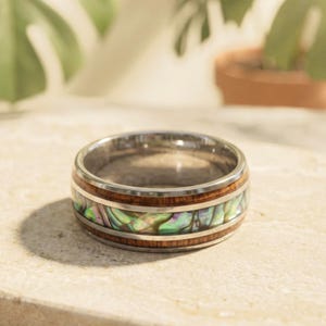 May include: A silver ring with a wooden band and iridescent abalone shell inlay. The ring has a polished finish and a wide band design. The abalone shell displays shades of green, blue, and purple. The ring is a piece of jewelry.